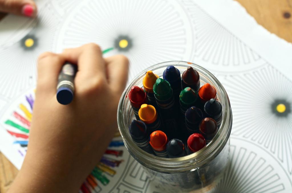 A picture of a child's hand coloring a page with a jar of crayons